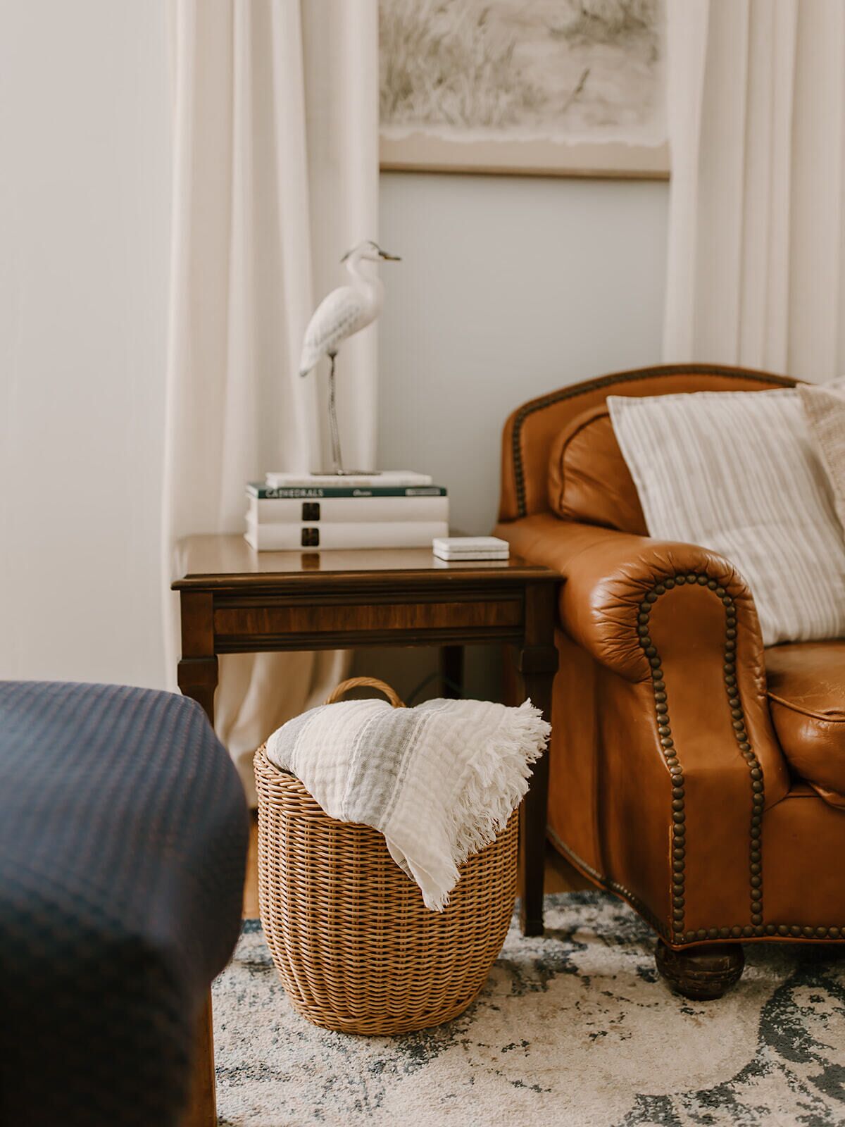 Une pièce chaleureuse avec un fauteuil en cuir marron, une table d’appoint en osier ornée de livres et d’un oiseau décoratif, et un panier tressé contenant un plaid blanc.