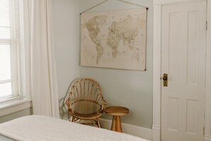 Um quarto com uma cama, uma cadeira de vime, um banquinho pequeno, um mapa-múndi na parede e uma porta.