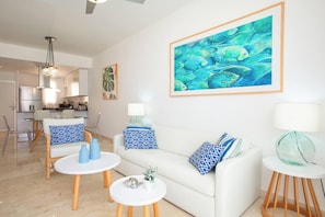 A modern living room with a white sofa, blue patterned cushions, a glass coffee table, and a dining area in the background. - Cana Rock Condos (Punta Cana)