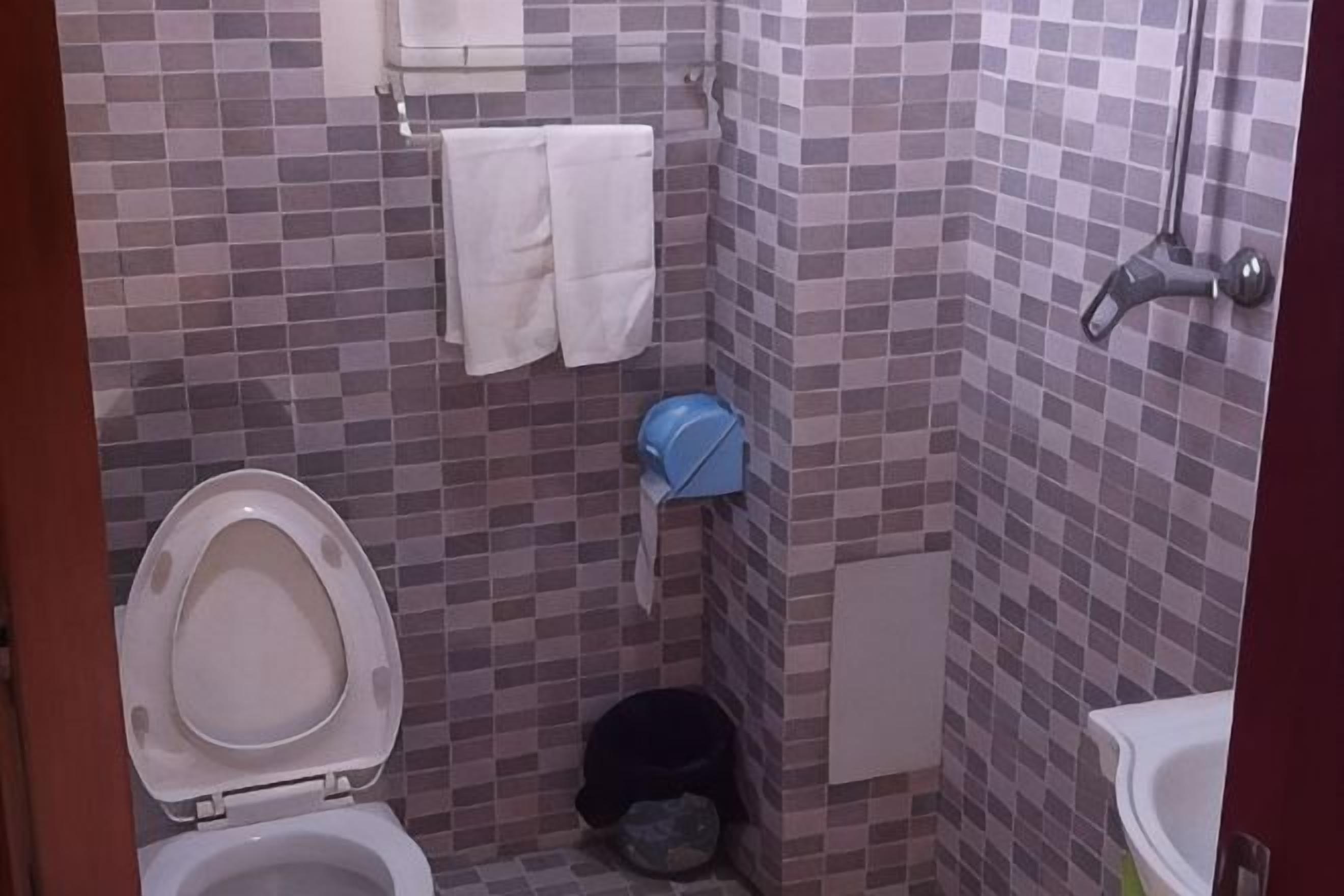 A small bathroom with a toilet, a sink, and a shower. There are towels hanging on the wall and a trash bin.