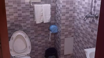 Une petite salle de bain avec des toilettes, un lavabo et une douche. Des serviettes sont accrochées au mur, et il y a une poubelle.