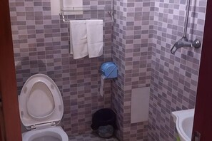 Un baño pequeño con inodoro, lavamanos y ducha. Hay toallas colgadas en la pared y un cubo de basura.