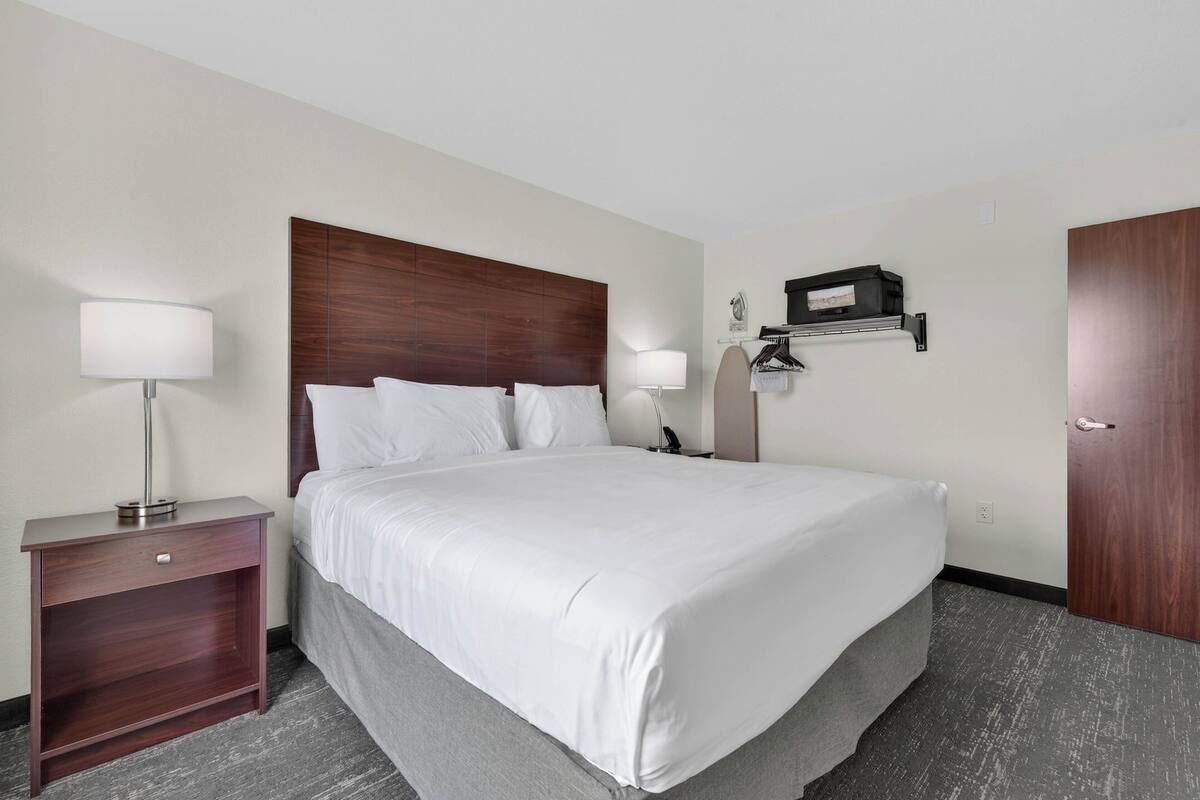A hotel room with a large bed, a nightstand with a lamp, a wall-mounted shelf with a TV, and a door.