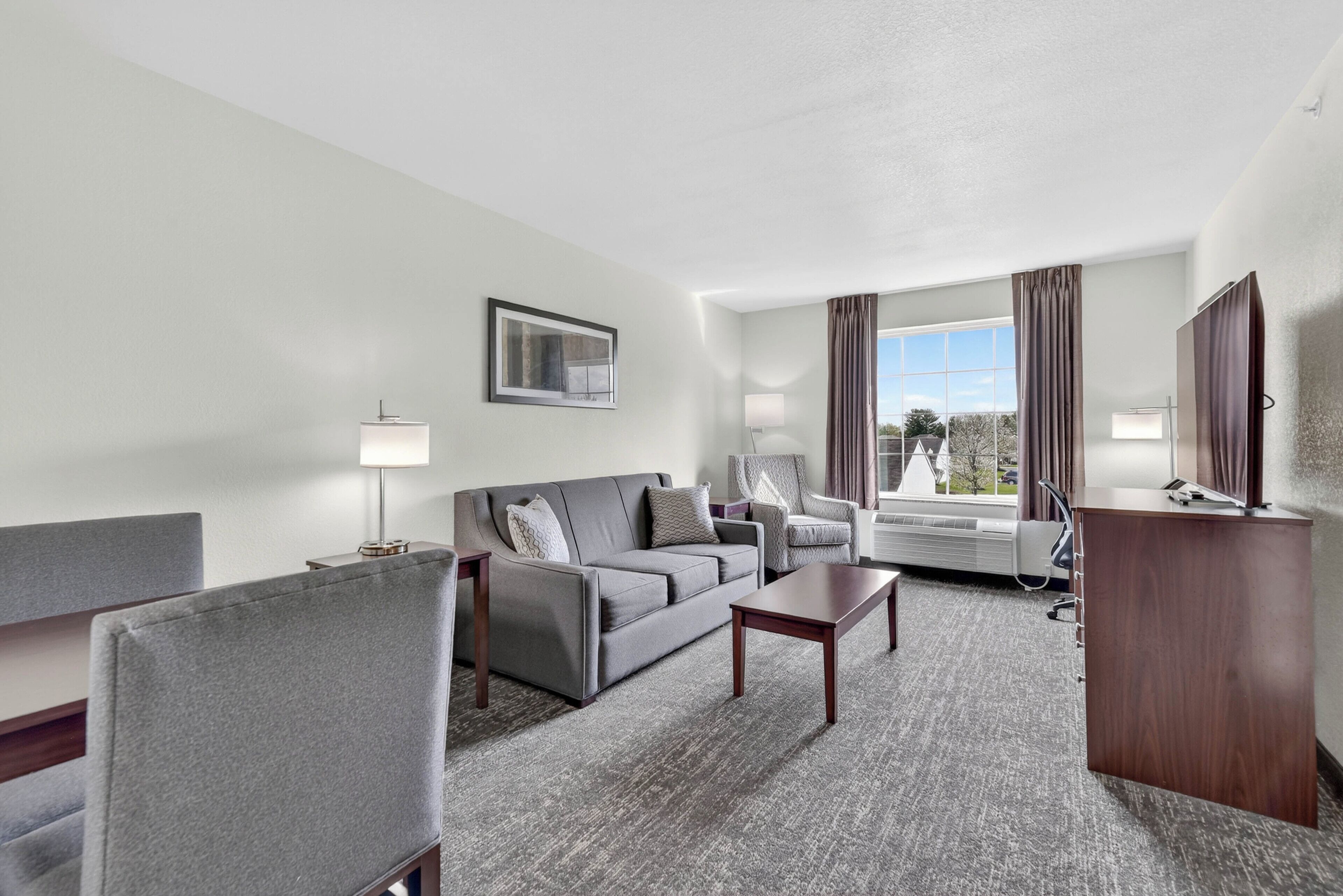 Une chambre d’hôtel avec un canapé gris, une petite table, une chaise, un bureau et une grande fenêtre avec des rideaux.