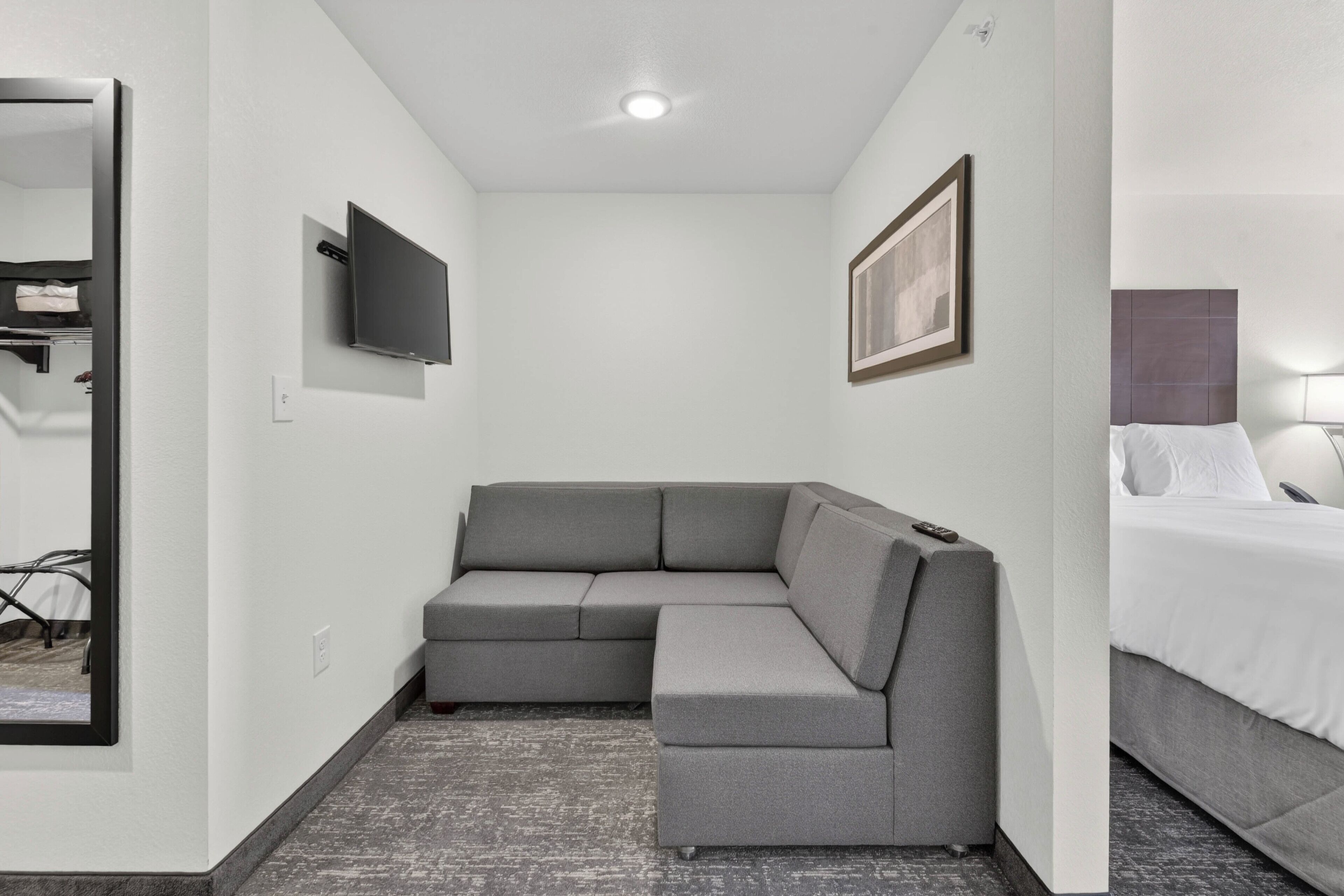 Een moderne hotelkamer met een grijze modulaire bank, een flatscreen televisie aan de muur, een bed met witte lakens en een ingelijst beeld boven het bed.