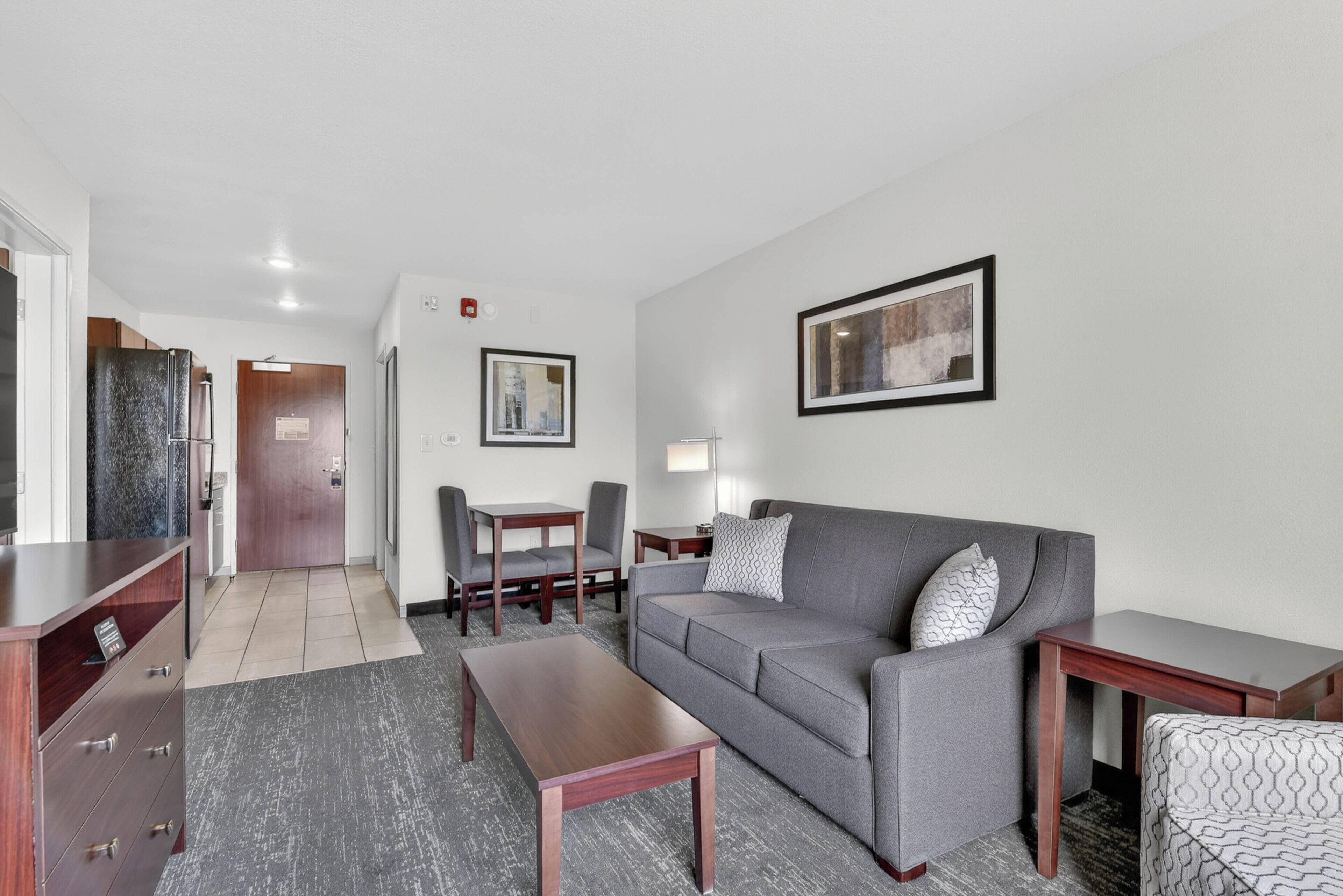 A modern living room with a grey sofa, wooden coffee table, and a patterned armchair. There is a dining area with a table and chairs, and a hallway leading to a door.