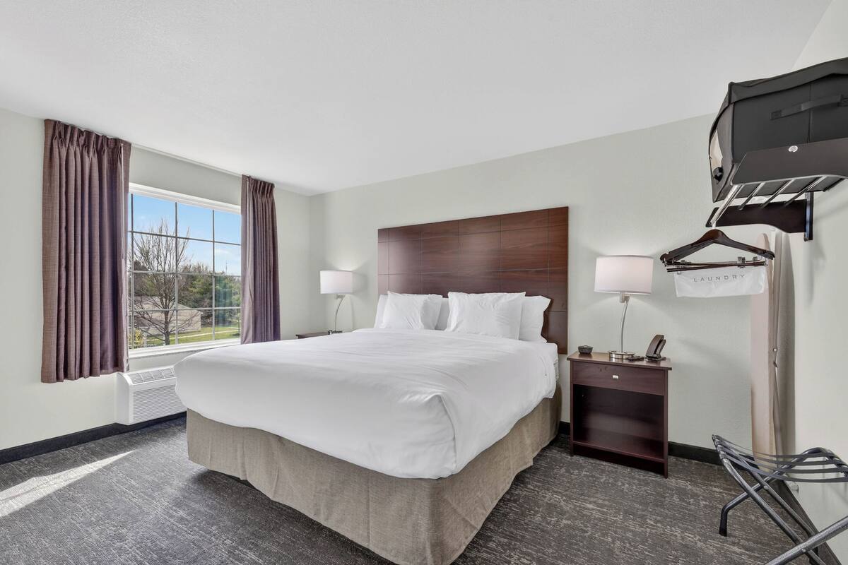A hotel room with a large bed, a nightstand with a lamp, a coat rack, and a view of the outdoors.