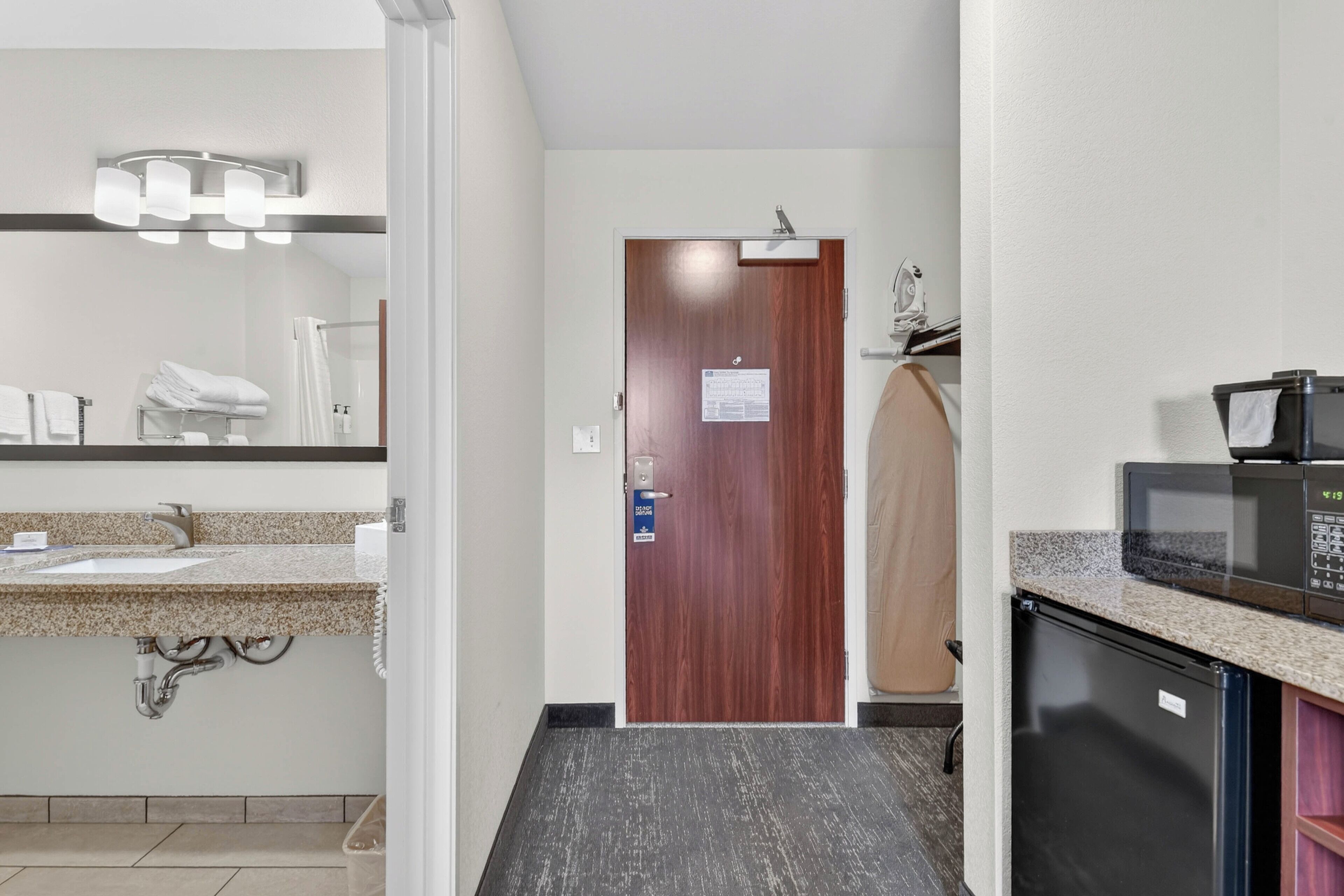 Een hotelbadkamer met een wastafel, spiegel, handdoekhouder en een deur met een bordje.