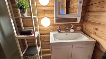 Ein Badezimmer einer Holzhütte mit einem weißen Waschtisch, einem Spiegel über dem Waschbecken und einem Regal mit Pflanze und gefalteten Handtüchern.