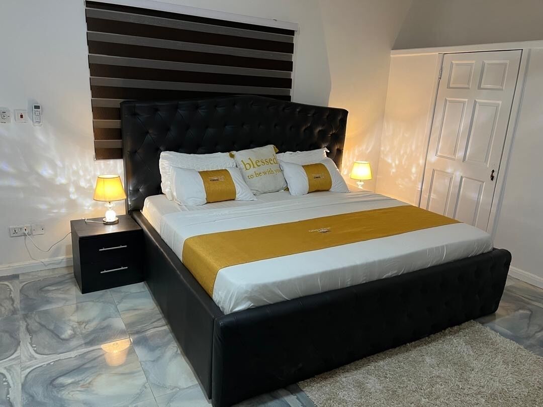 Una camera da letto con una testiera imbottita nera, un letto con lenzuola bianche e gialle, due abat-jour, un comodino con cassetti e un pavimento con decoro marmorizzato.