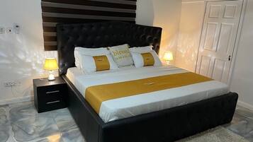 Una camera da letto con una testiera imbottita nera, un letto con lenzuola bianche e gialle, due abat-jour, un comodino con cassetti e un pavimento con decoro marmorizzato.
