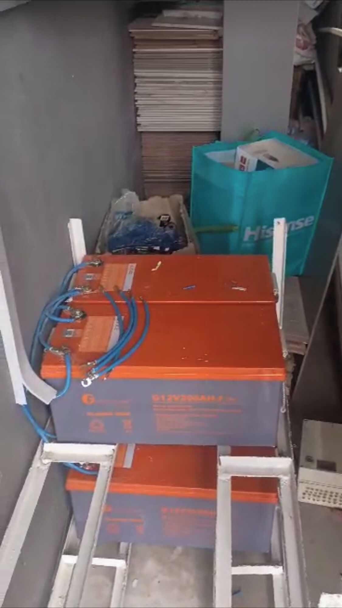 A narrow hallway with a metal cart carrying orange and blue battery boxes. A teal shopping bag is visible in the background.