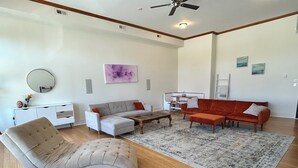 Ein geräumiges Wohnzimmer mit einer grauen Eckcouch, einem weißen Sessel und einem roten Sofa. Ein weißer Konsolentisch mit Spiegel und Blumenvase steht dort. Ein Deckenventilator ist sichtbar, und der Boden ist mit Holzdielen belegt.