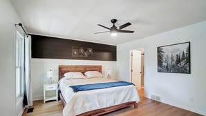 Une chambre à coucher avec un lit en bois, du linge de lit blanc, un ventilateur de plafond, une table de chevet avec une lampe et un tableau encadré au mur.