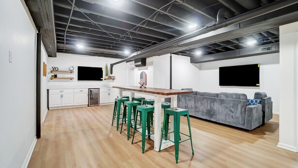 Une cuisine moderne avec des armoires blanches, un comptoir en bois et un four encastré. On y trouve des tabourets de bar verts et un canapé gris dans l’espace repas.