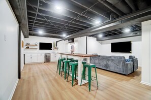 Una cocina moderna con armarios blancos, encimera de madera y horno integrado. Hay taburetes altos verdes y un sofá gris en la zona de comedor.