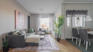 A modern living room with a grey sofa, a coffee table, and a dining area with a table and chairs. There is a large window with curtains, a potted plant, and a painting on the wall. - Homelike 1BR Apt. with Sauna & Parking (Kuopio)