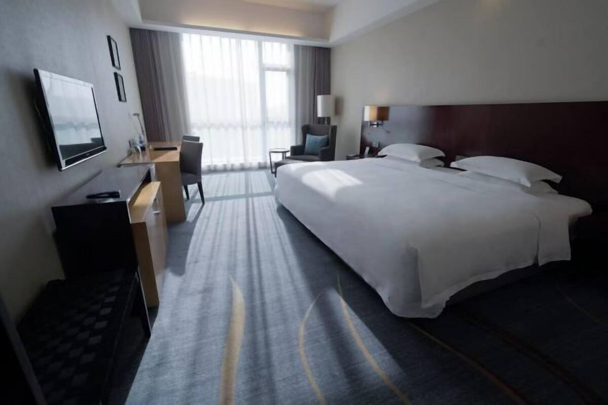 A hotel room with a large bed, a desk with a chair, a TV mounted on the wall, and a window with curtains.