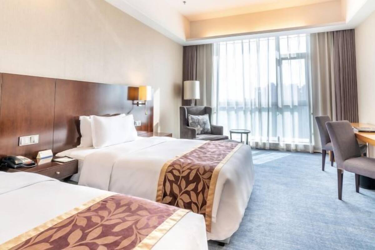 A hotel room with two beds, a desk, a chair, a lamp, and a view of the city through large windows.