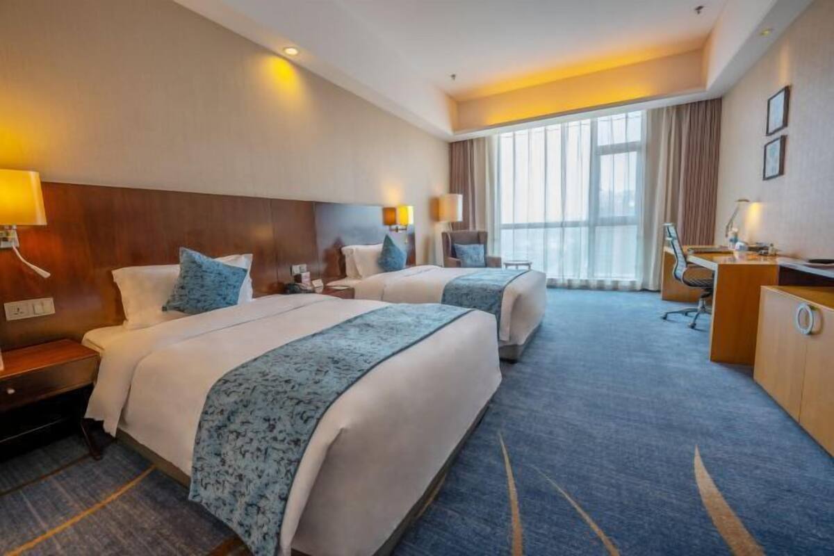 A hotel room with two beds, a desk, a chair, and a large window.