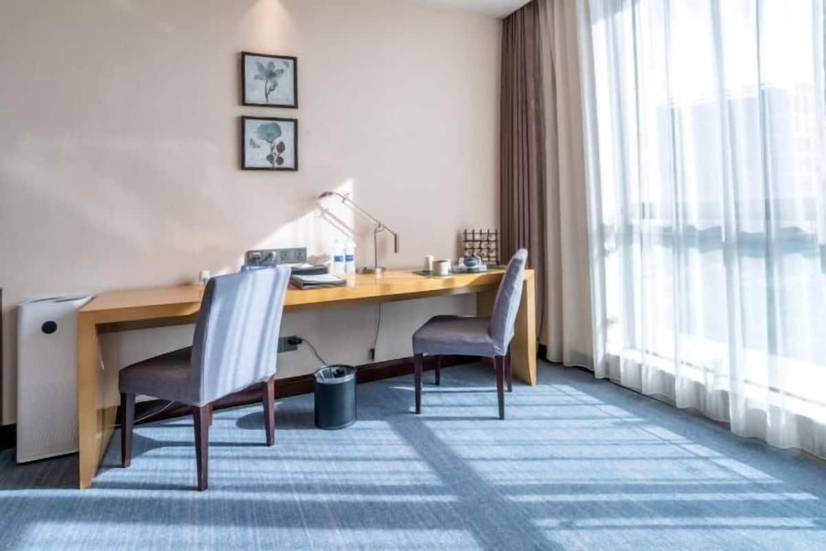 A hotel room with a wooden desk, two chairs, a lamp, a small trash bin, and a window with sheer curtains.