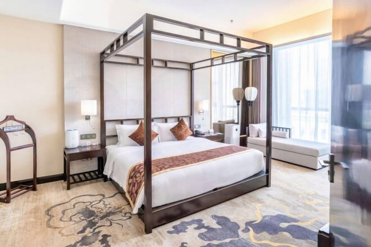 A four-poster bed with a wooden frame, a bedside table with a lamp, a sofa, and a view of the city through large windows.