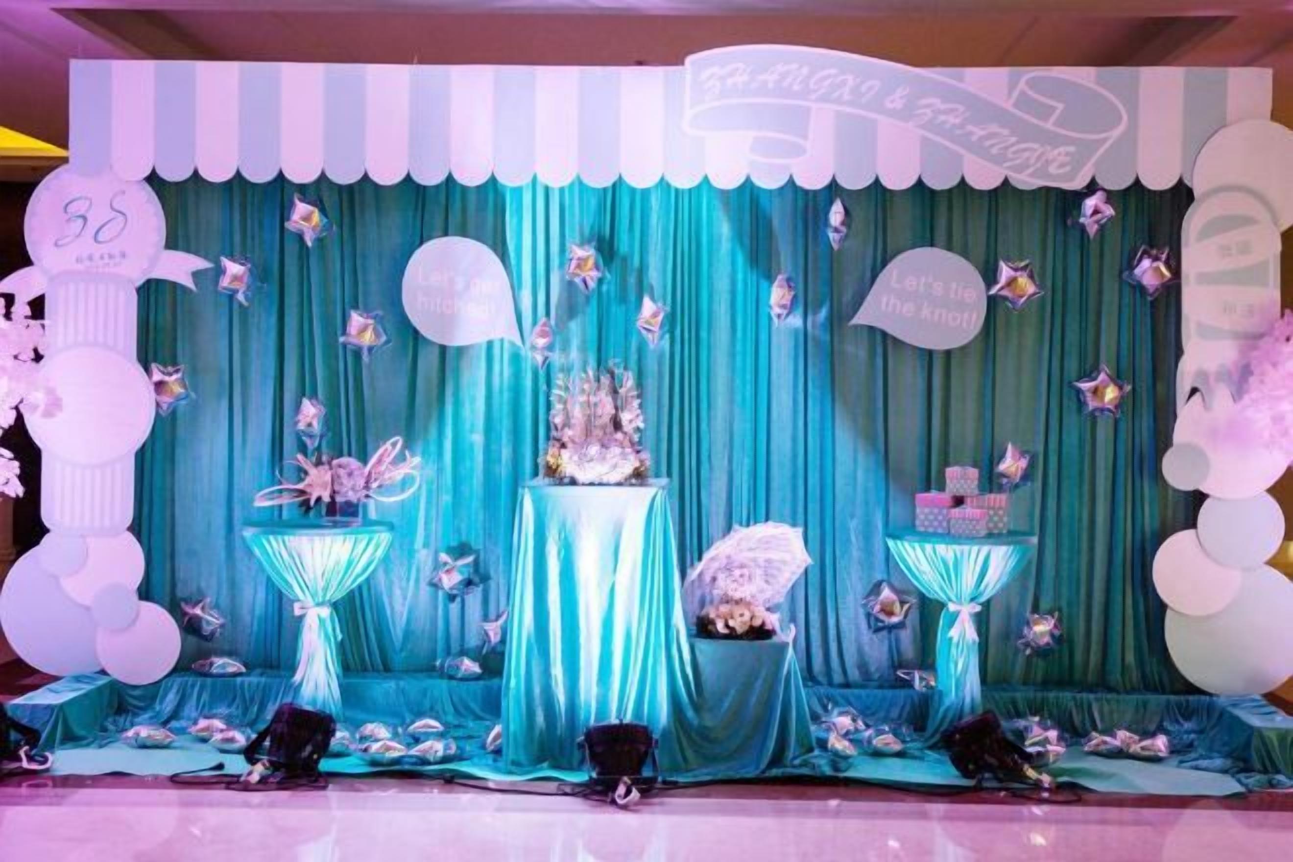 A stage with a teal curtain backdrop, a central floral arrangement, and decorative elements like balloons and a sign.