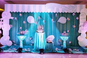 Un escenario con un telón de fondo color aguamarina, un arreglo floral central y elementos decorativos como globos y un cartel.