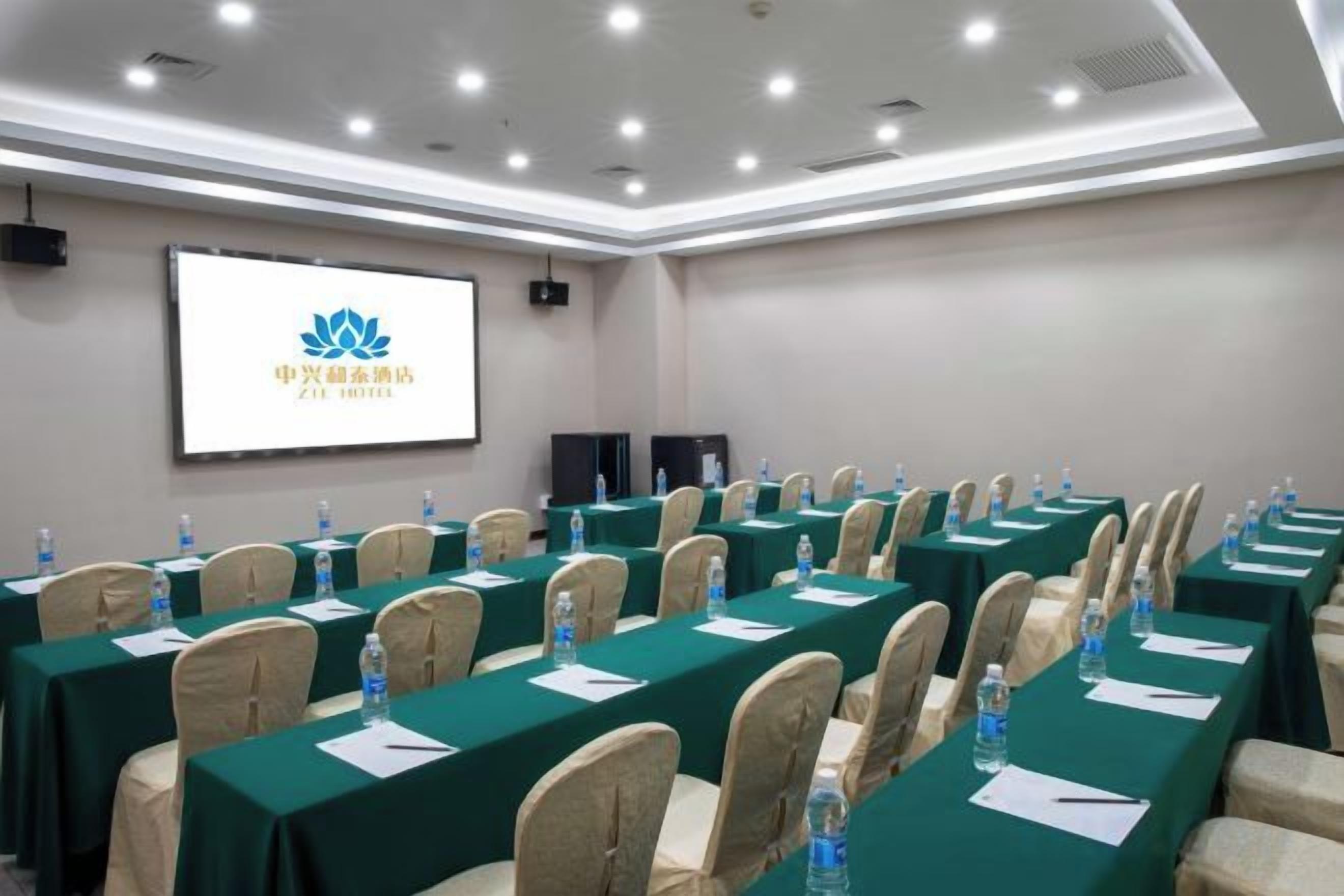 A conference room with rows of tables, chairs, and a projection screen displaying a logo.