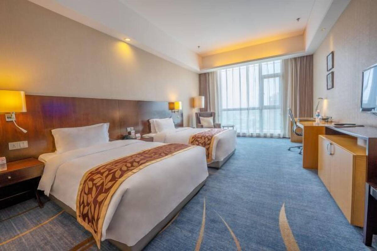 A hotel room with two beds, a desk, a TV, and a large window.