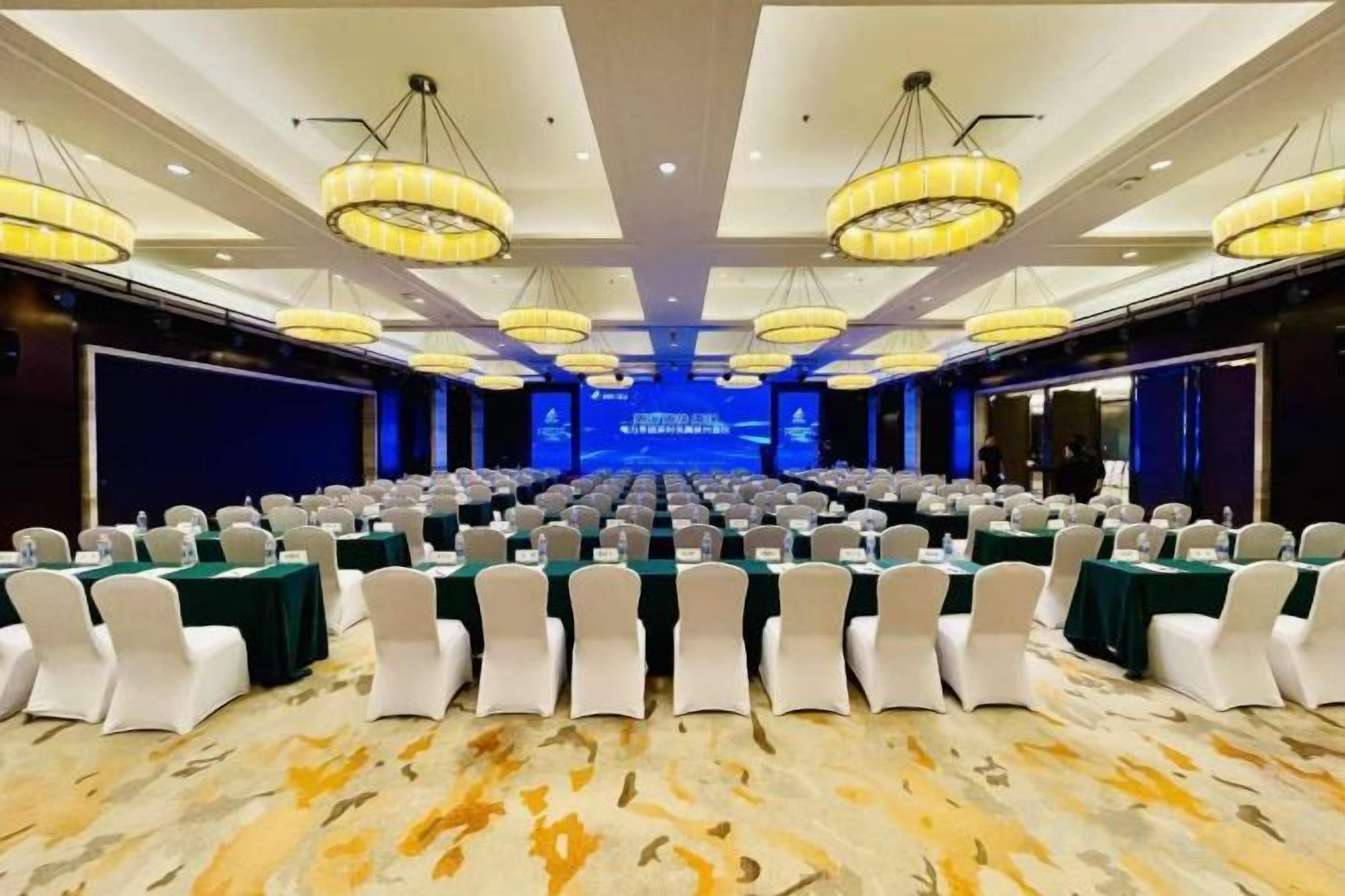 Un salón de conferencias con filas de mesas y sillas, grandes candelabros y un escenario al frente.