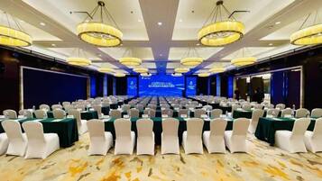 A conference room with rows of tables and chairs, large chandeliers, and a stage at the front.
