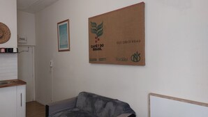 A room with a framed poster on the wall, a telephone, a shelf with kitchen items, and a grey sofa. - The budget studio. (Limoges)