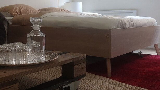 A bedroom with a bed, a wooden nightstand, a glass decanter, and a guitar on the wall. - The budget studio. (Limoges)
