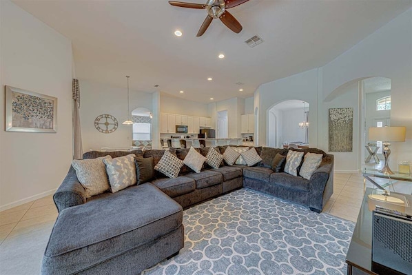 A spacious living area with a large sectional sofa, a ceiling fan, recessed lighting, and a kitchen in the background.