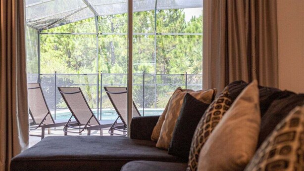 A dark-colored sofa with patterned cushions, a glass door leading to a balcony with two chairs and a pool view.