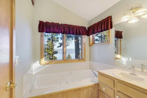 Un baño con una bañera grande, un lavabo y un espejo. Hay ventanas con cortinas, y se ve una vista de árboles y montañas.