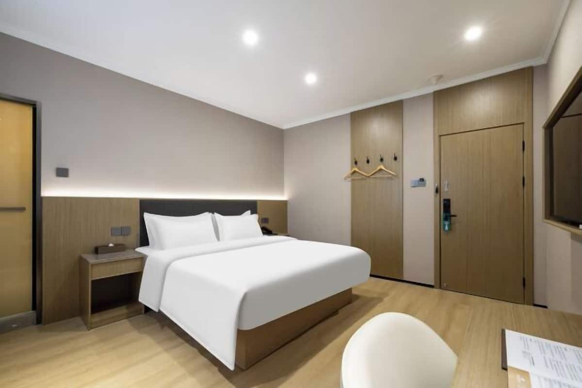 A modern hotel room with a bed, bedside table, wardrobe, and a desk with a chair.