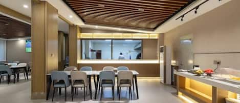A modern dining area with a long table and chairs, a counter with food, and a view of a kitchen area.