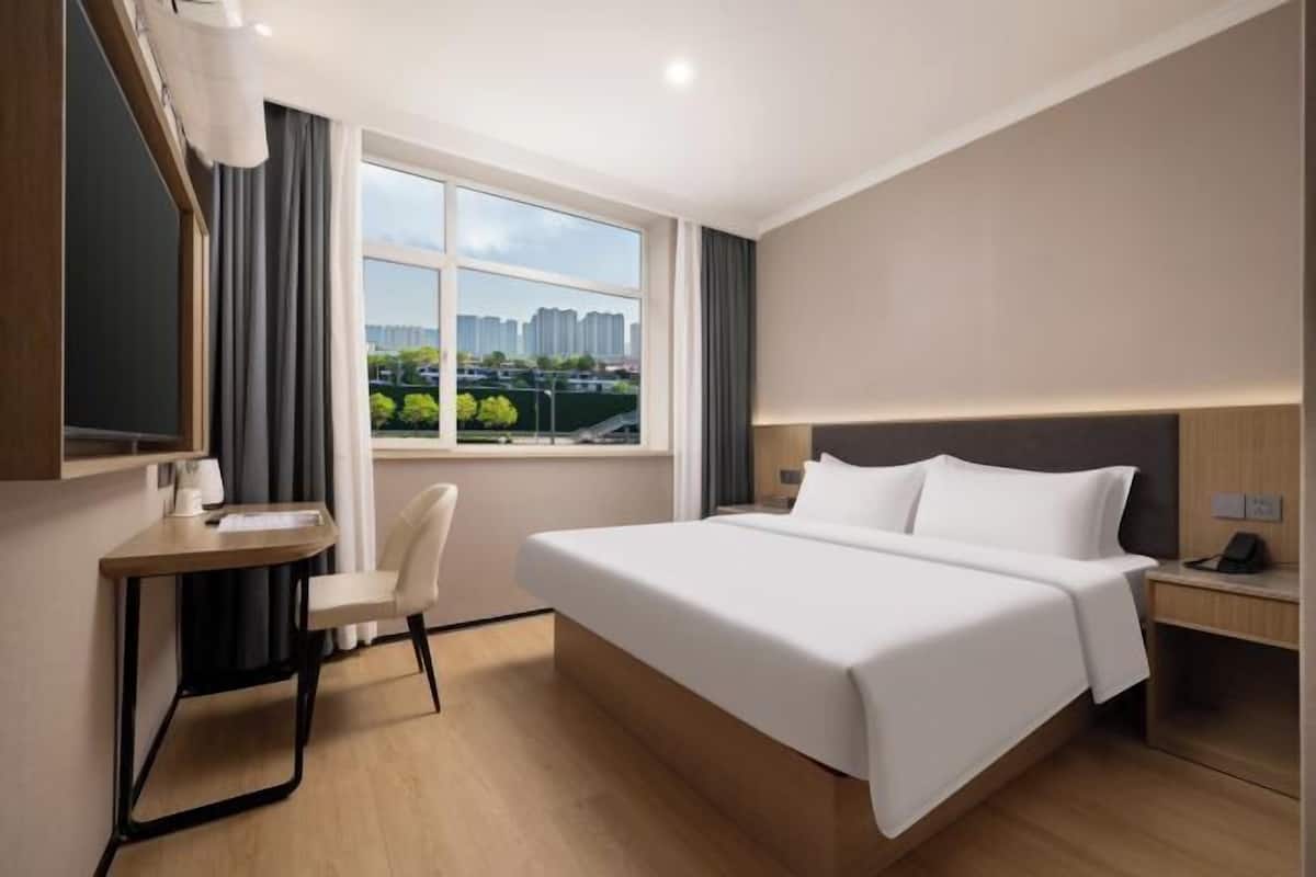 A hotel room with a large bed, a desk, a chair, a TV, and a window with a city view.
