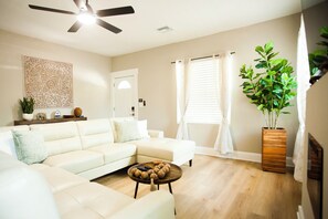 Una sala de estar con un sofá modular, una mesa de centro redonda con adornos, un ventilador de techo y una planta grande en maceta.