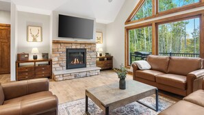 A spacious living room with a stone fireplace, leather sofas, a flat-screen TV, and large windows offering a view of the outdoors. - Riverfront Chalet on 5 acres~GameRoom~Spa~Gym~Firepit~Theater~2min to CrystalMtn (Thompsonville)