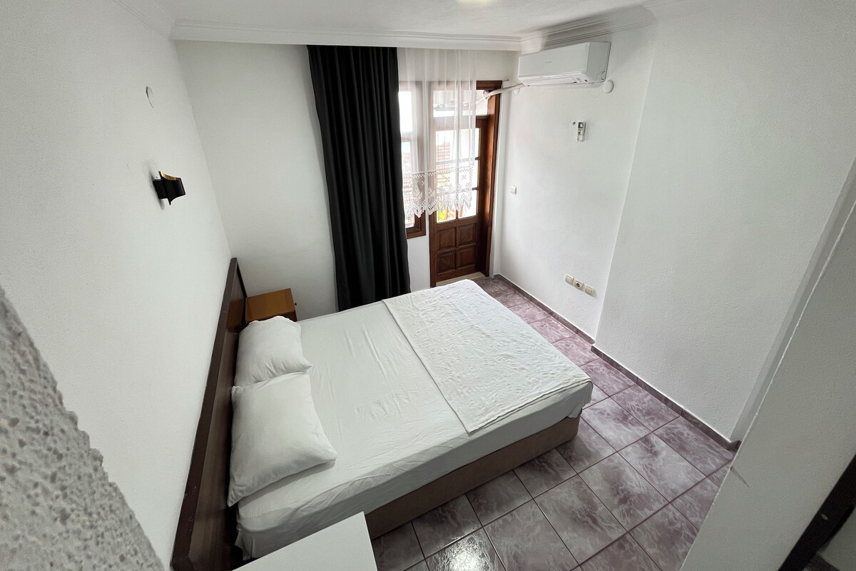 Una habitación pequeña y limpia con cama, mesita de noche, ventana con cortinas y aire acondicionado.