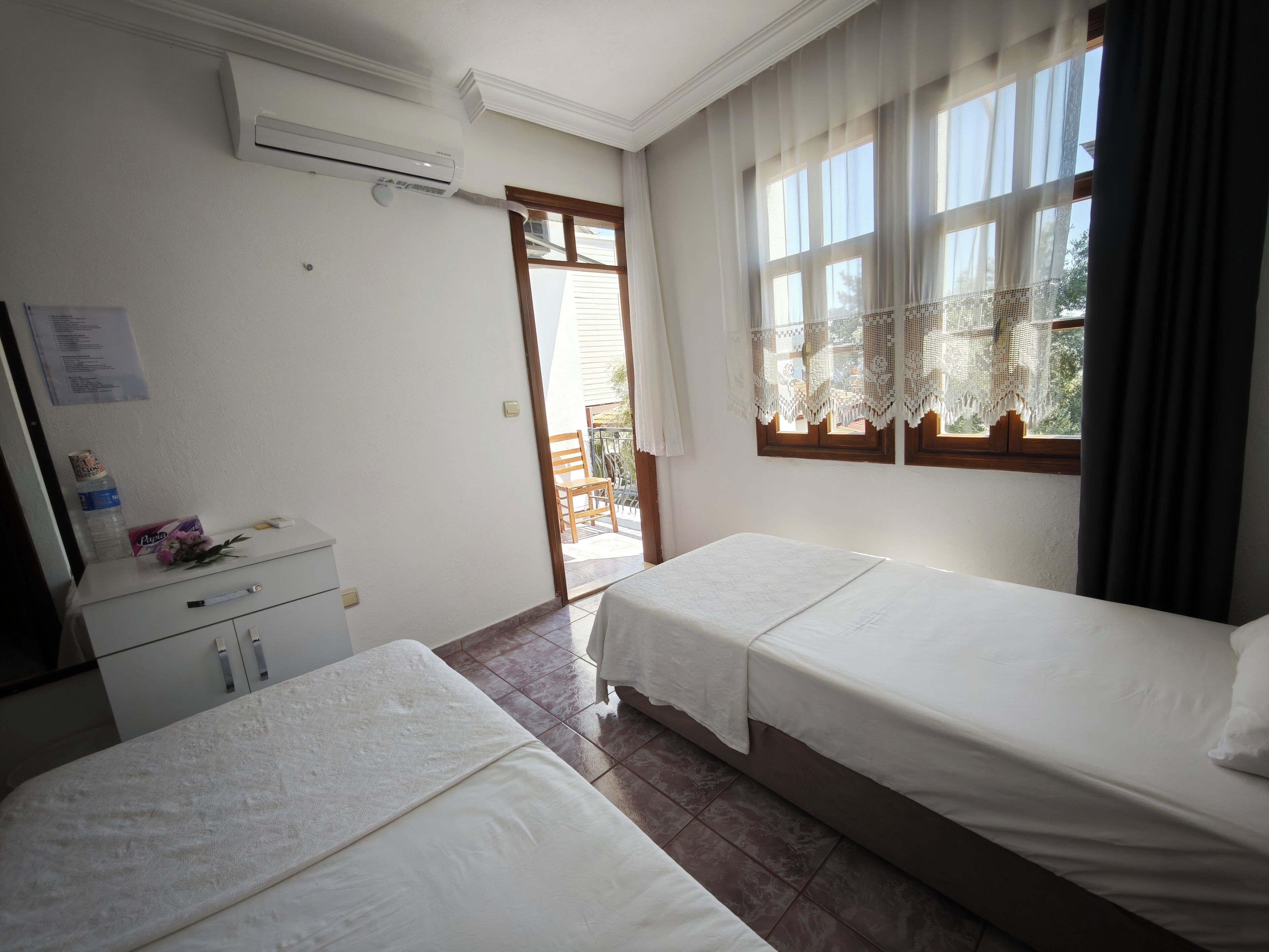 A hotel room with two beds, a desk, a chair, and a window with lace curtains.