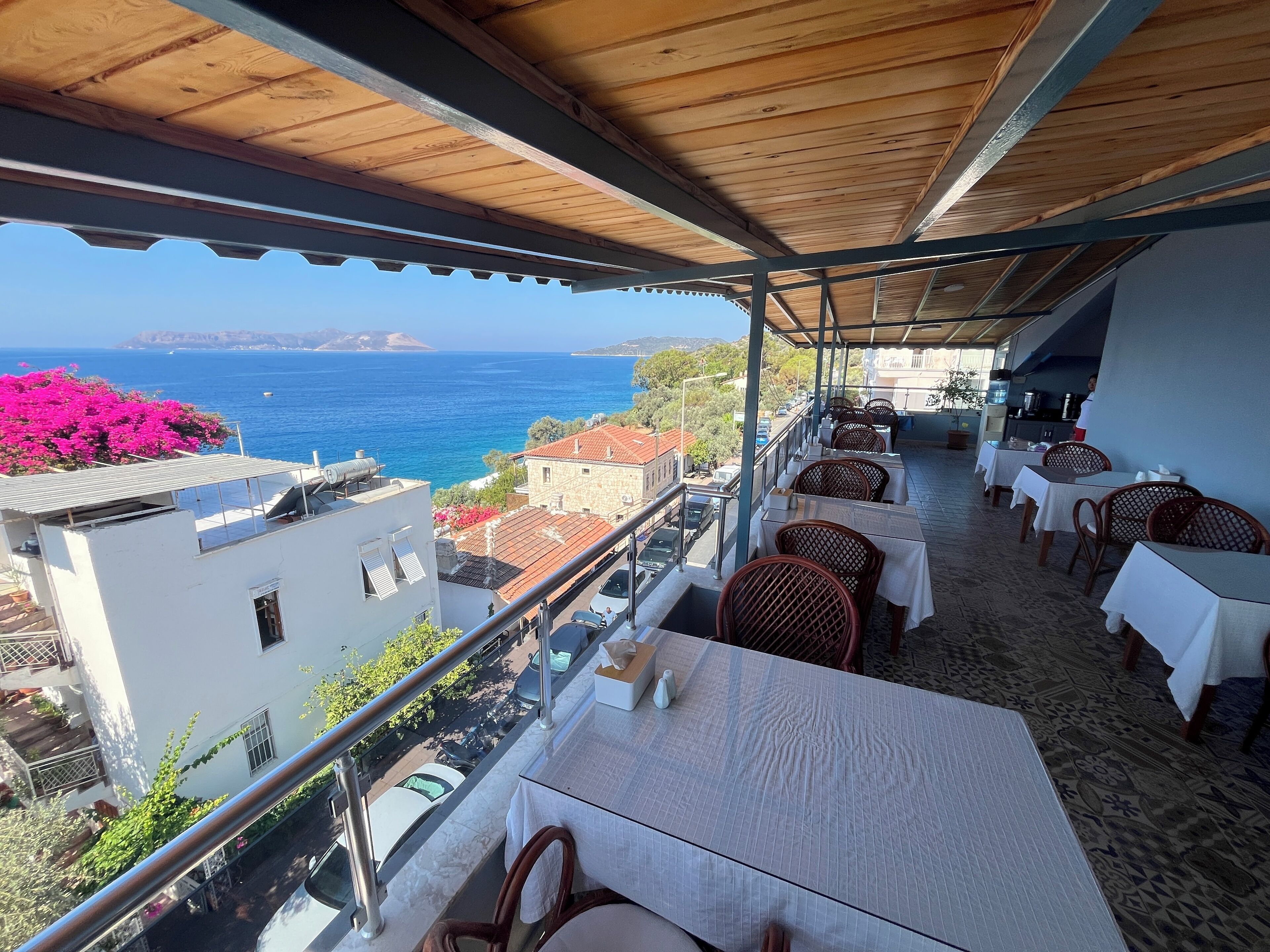 A terrace with tables and chairs, overlooking a coastal view with buildings and a clear sky.