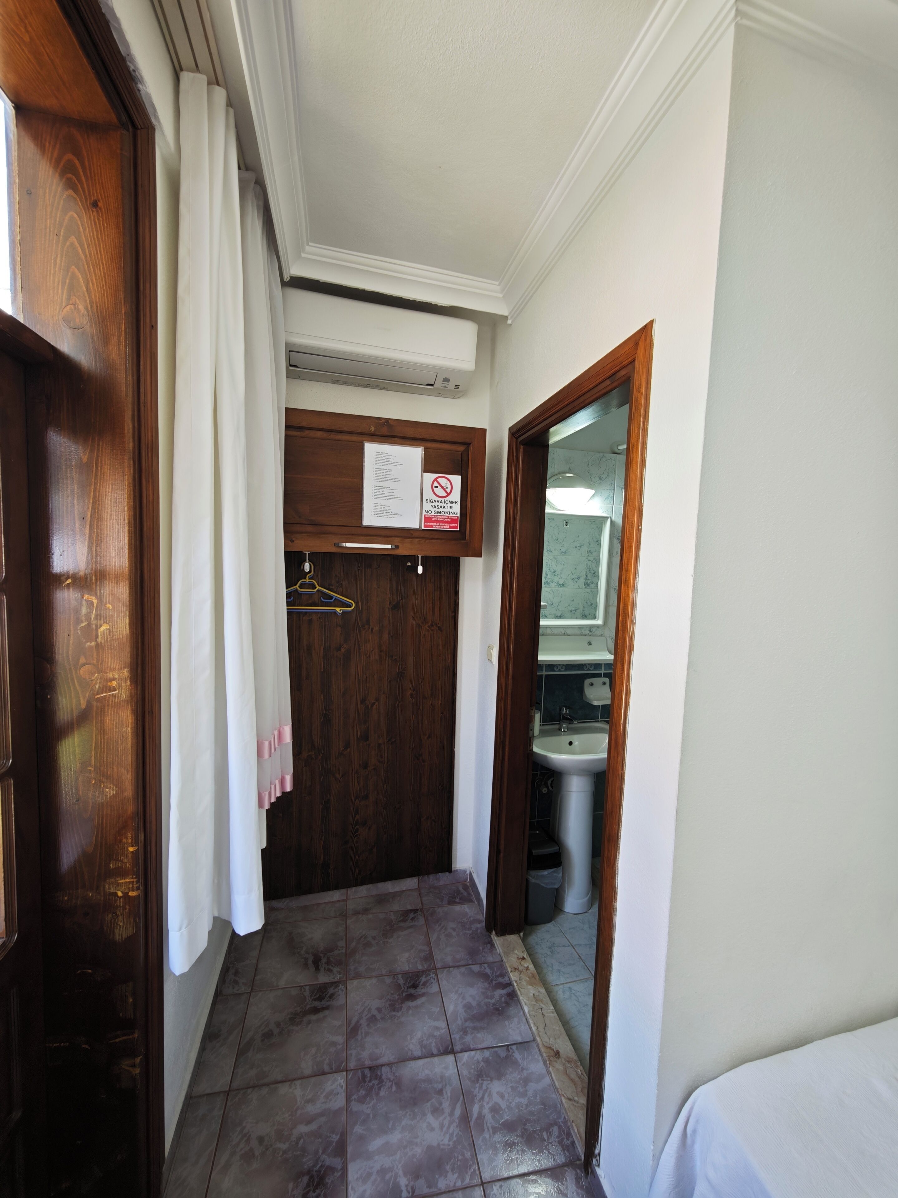 Un corridoio con una porta in legno, tende bianche, un cartello con divieto di fumare e vista su un bagno con lavandino e specchio.