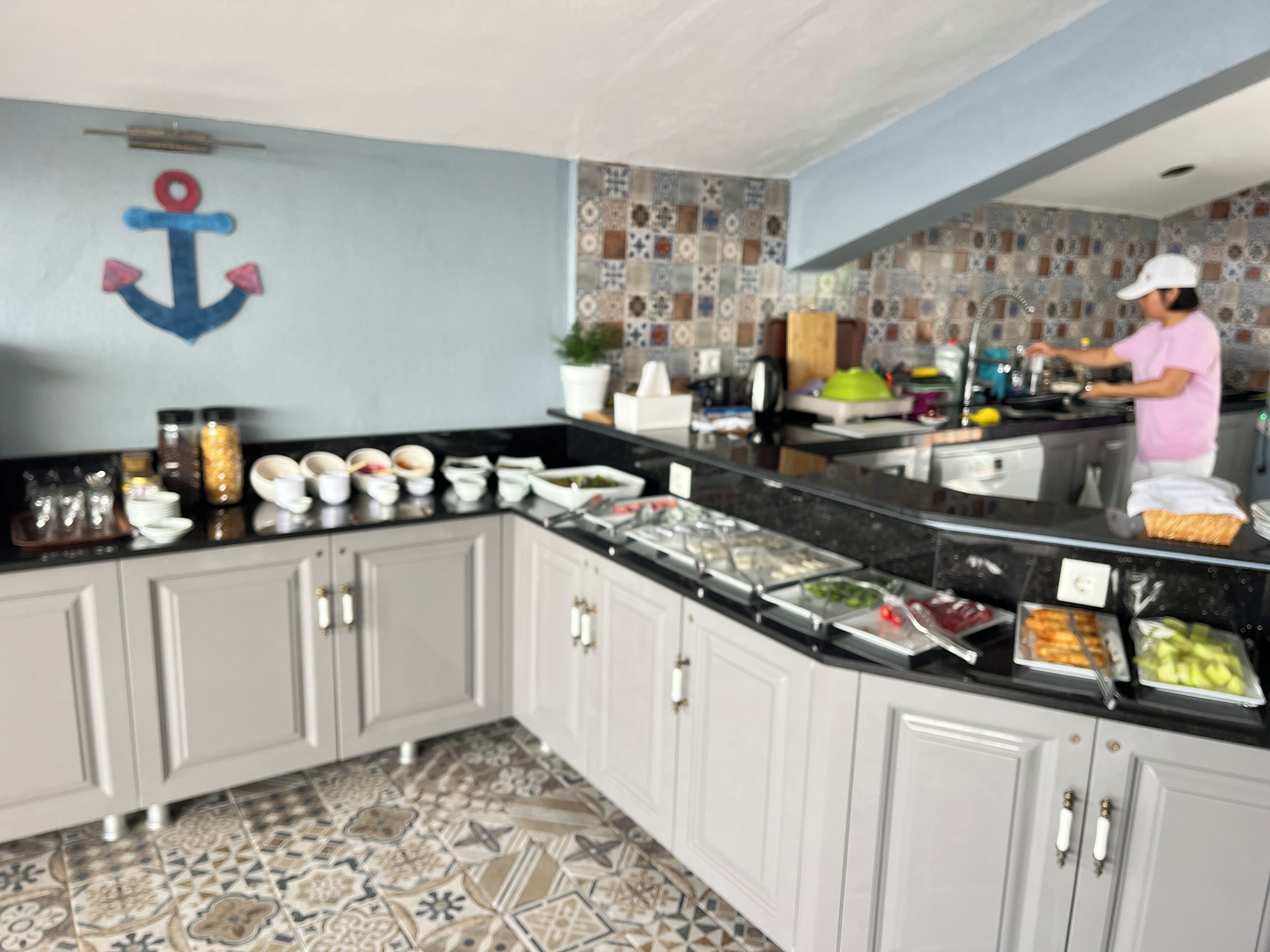 Una cocina con salpicadero de azulejos, un mostrador tipo buffet con diferentes alimentos, y una persona preparando comida.