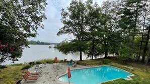 A swimming pool with stone tiles, surrounded by grass and trees, with a view of a lake and a dock in the background. - A Waterfront Retreat with pier and pool.  Spacious 4 bedrooms near Chestertown. (Chestertown)