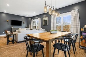 Una sala de estar moderna con sofá, mesa de comedor, sillas, televisor de pantalla plana y un amplio ventanal con cortinas.