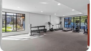 Un gimnasio con equipo cardiovascular, mancuernas y amplias ventanas.